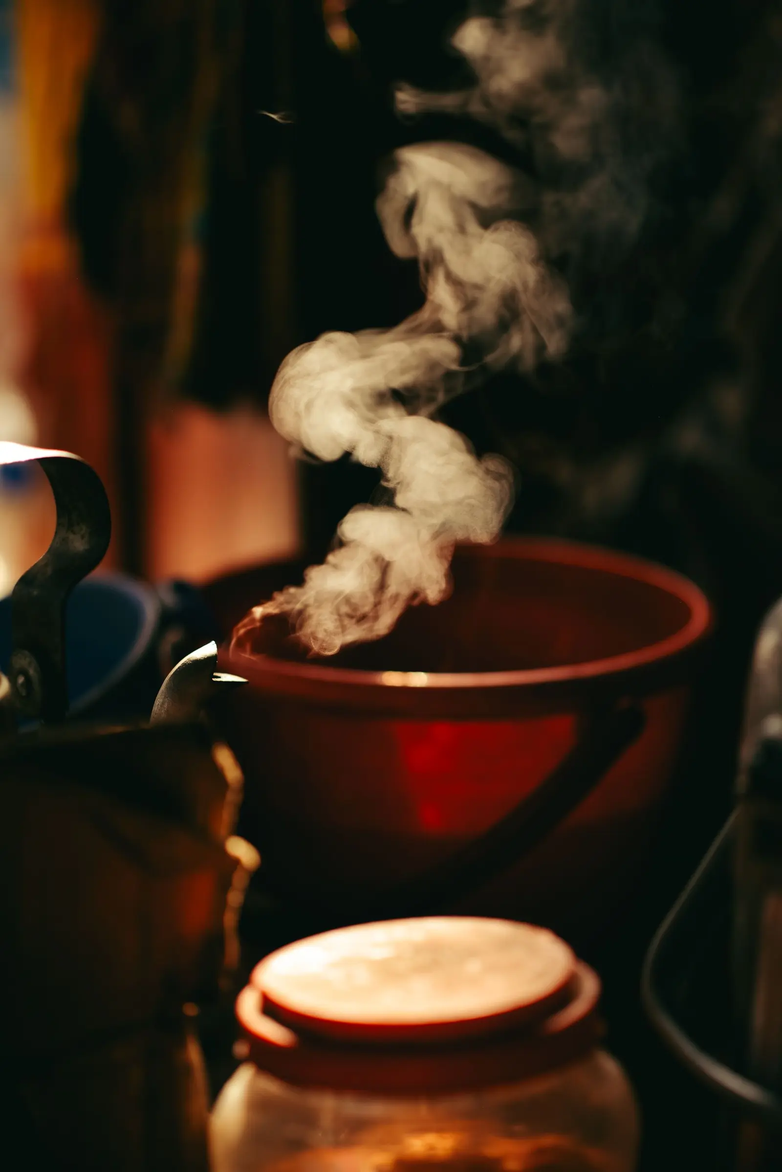 Steam rising from a kettle beside a glowing pot in a dark kitchen