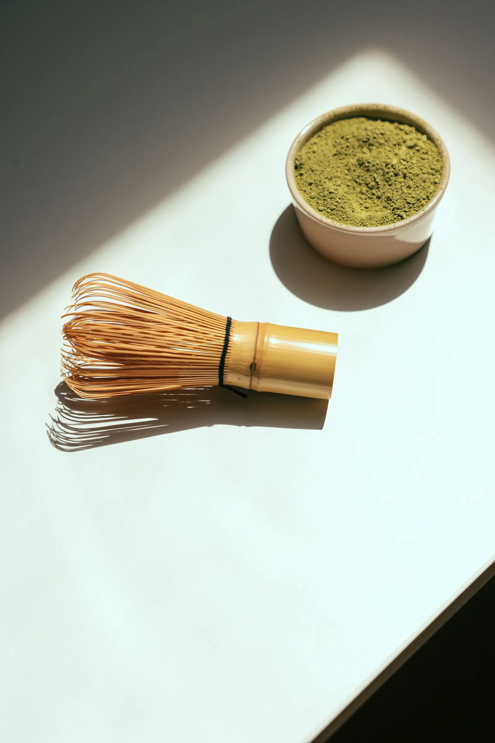 Small bowl of matcha powder beside a bamboo whisk in bright sunlight