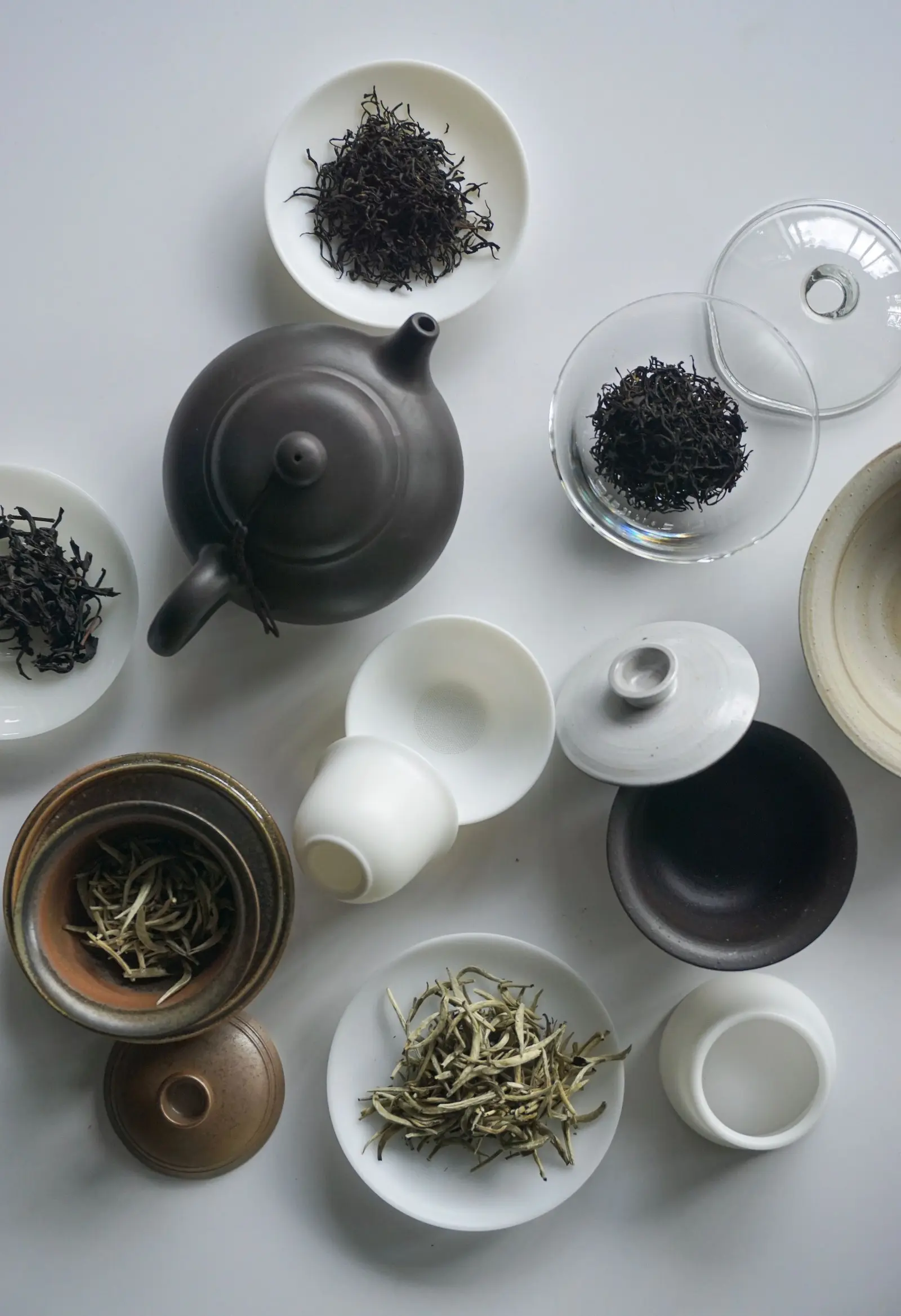 Flat lay of a dark teapot, small bowls of loose tea and white tea cups