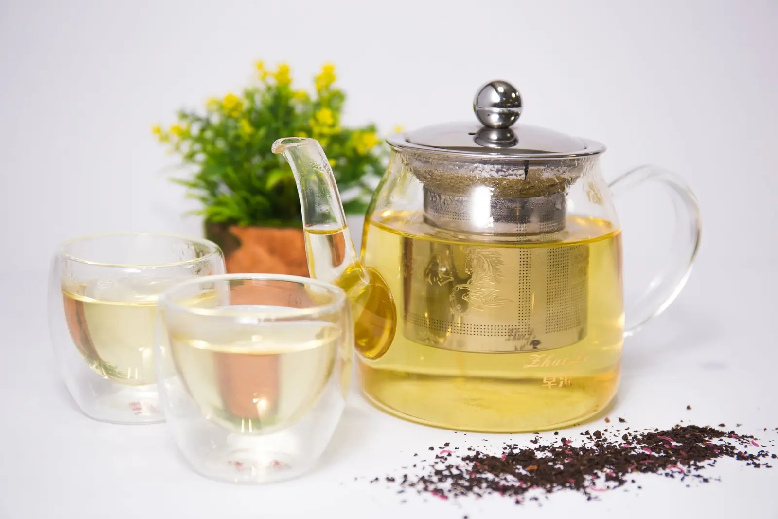Glass teapot with a steel infuser, two double-walled cups and loose tea leaves on a white table