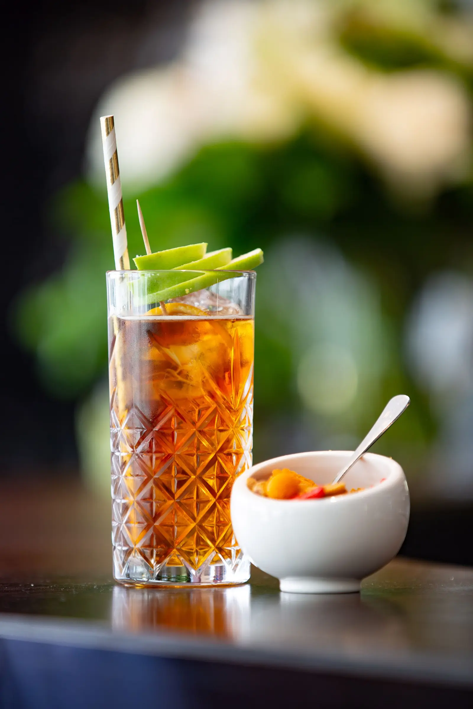 Tall glass of iced tea with a straw and a small bowl on a dark table