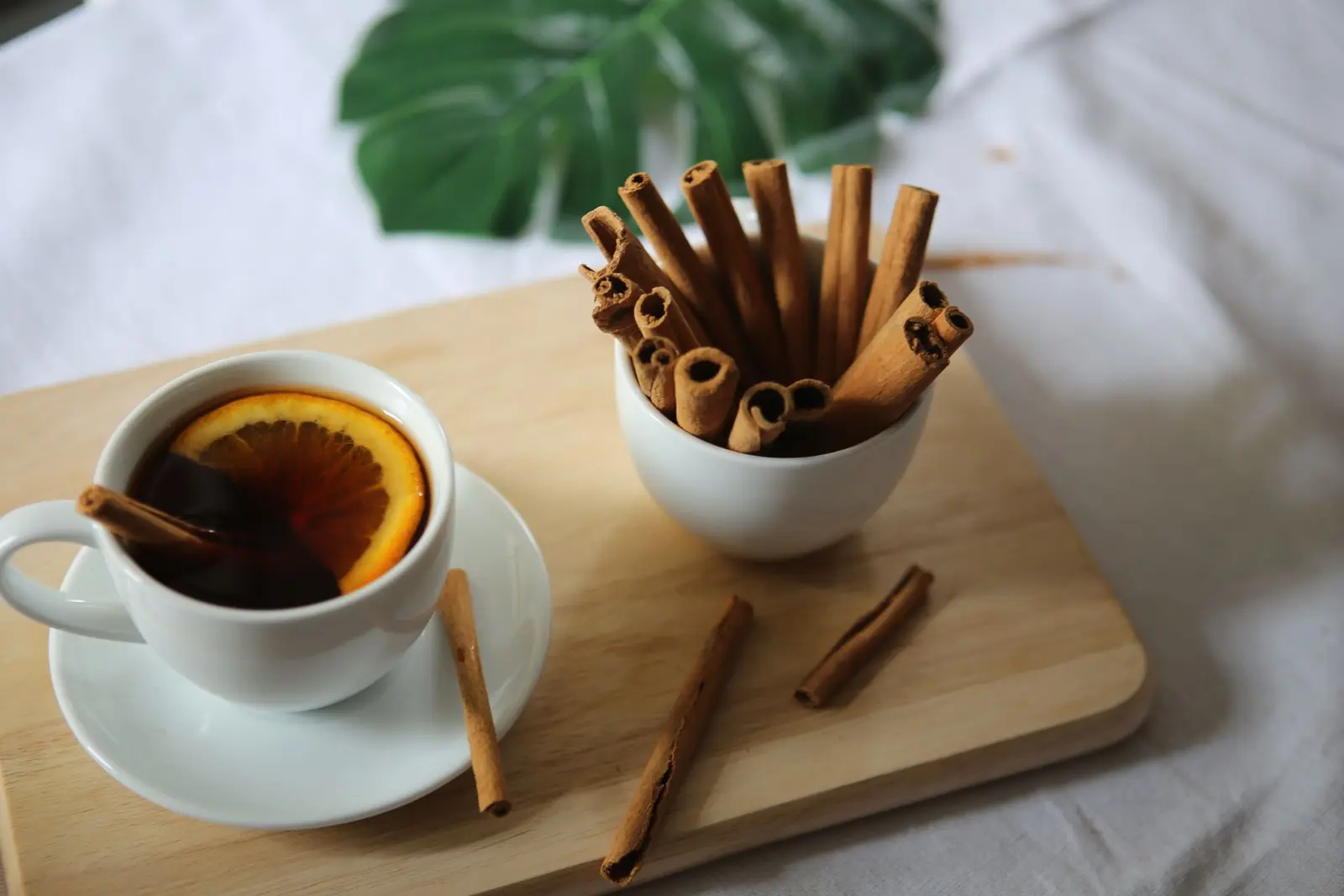 Cup of spiced tea with an orange slice next to a bowl of cinnamon sticks on a wooden board