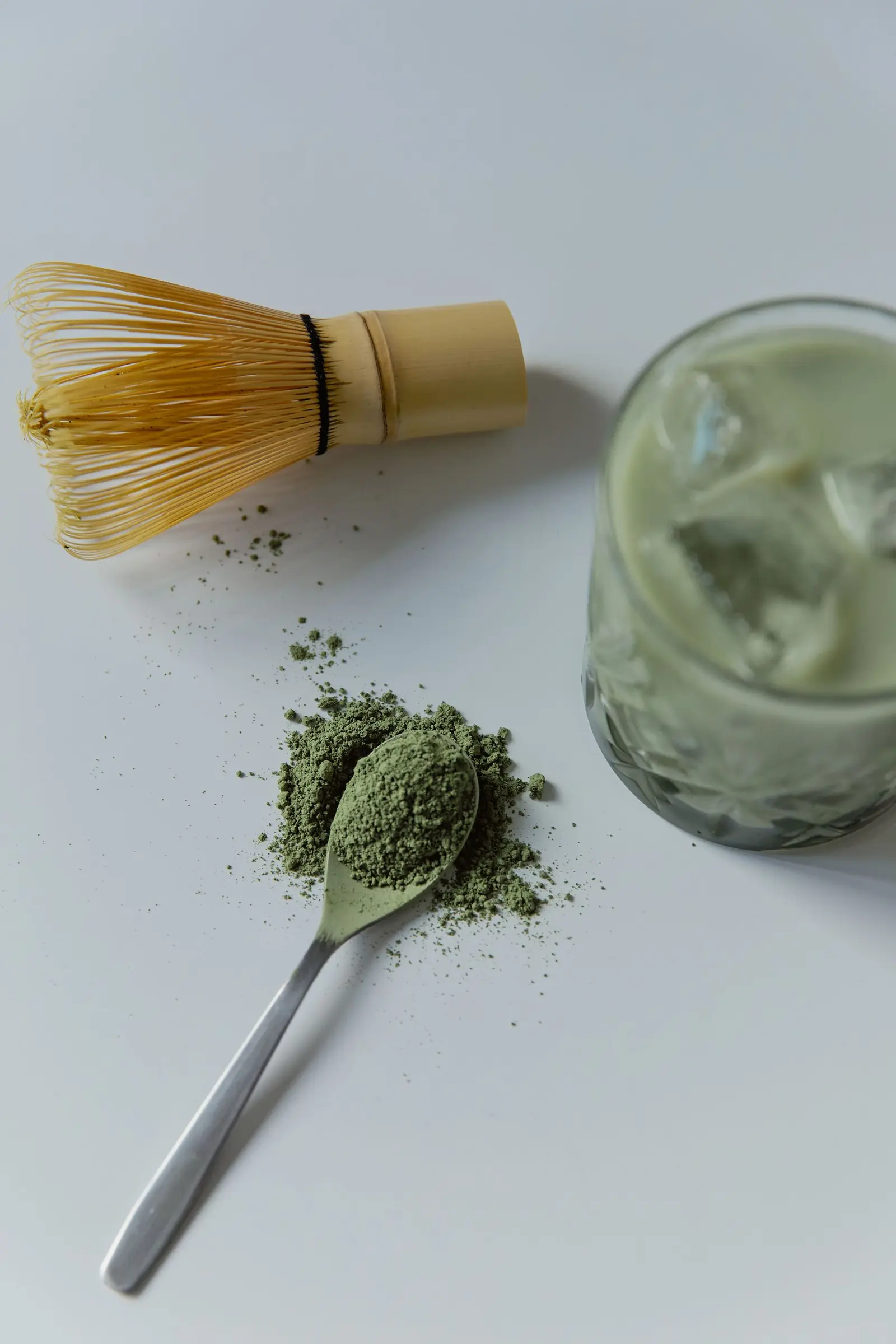 Bamboo whisk, a spoon of matcha powder and a glass of iced matcha on a pale surface
