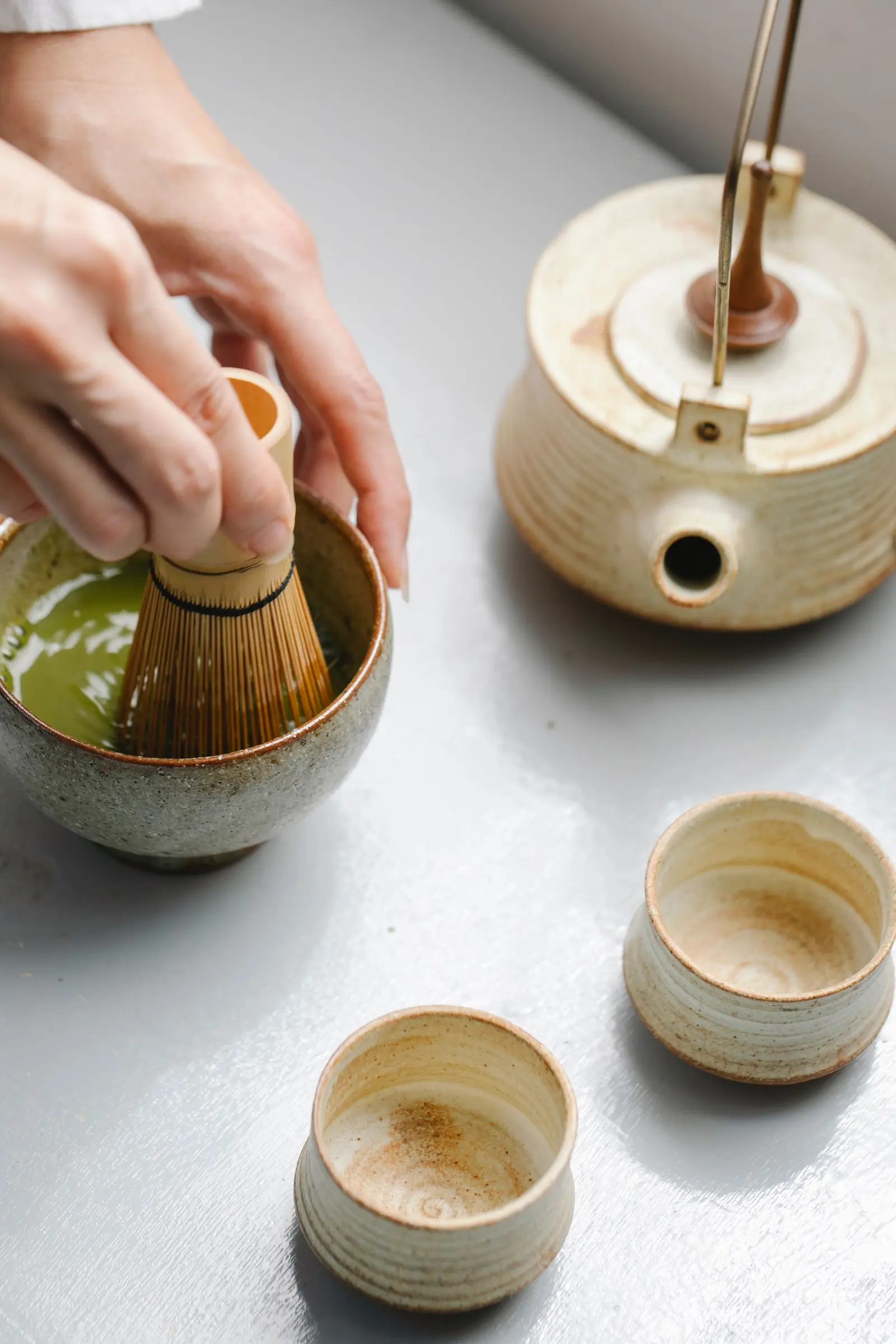 Hands whisking matcha in a bowl beside a small teapot and tea cups