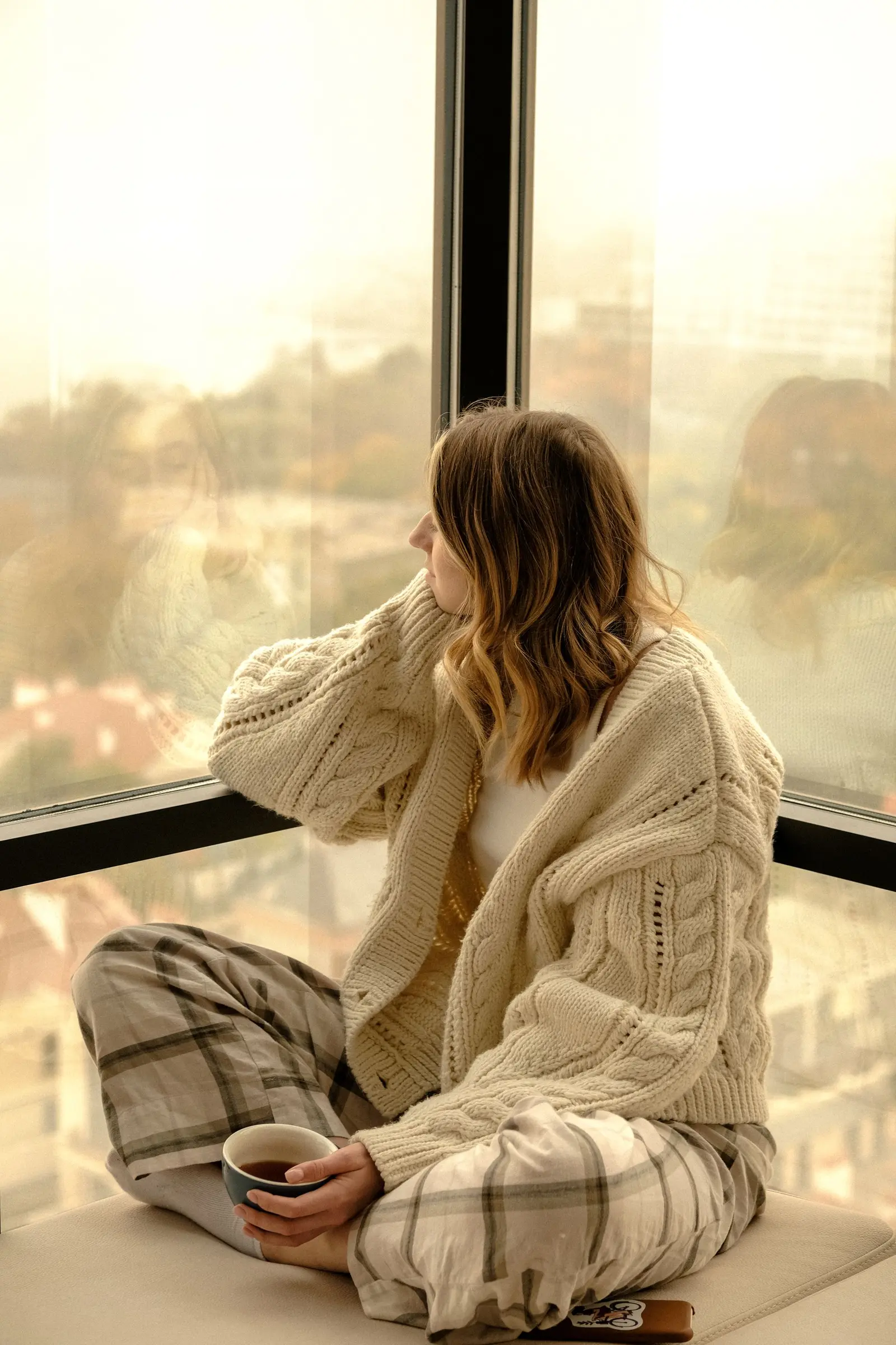 Woman in a cozy sweater sitting by a window holding a cup of tea